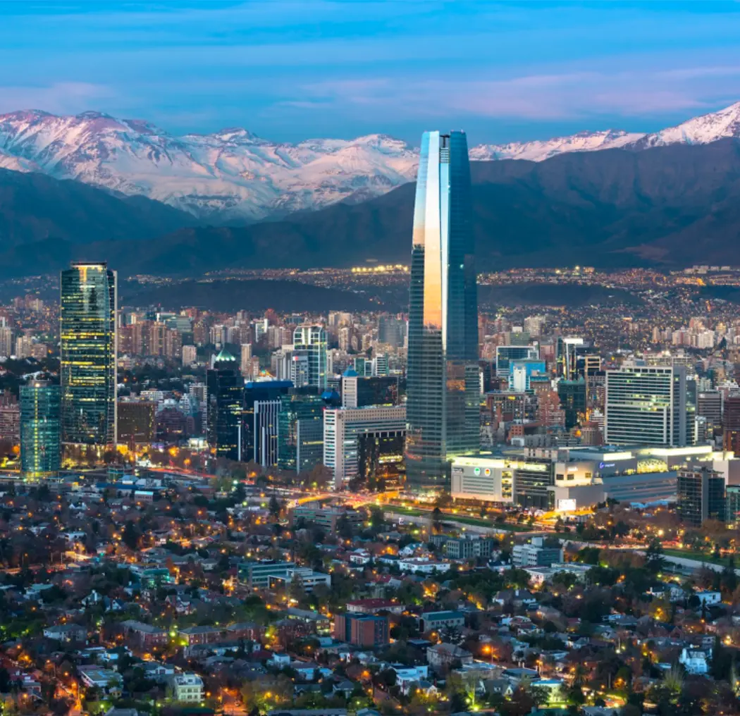 Aerial view of Santiago, Chile, featuring the Gran Torre Santiago amidst the city skyline with snow-capped Andes Mountains in the background during twilight.