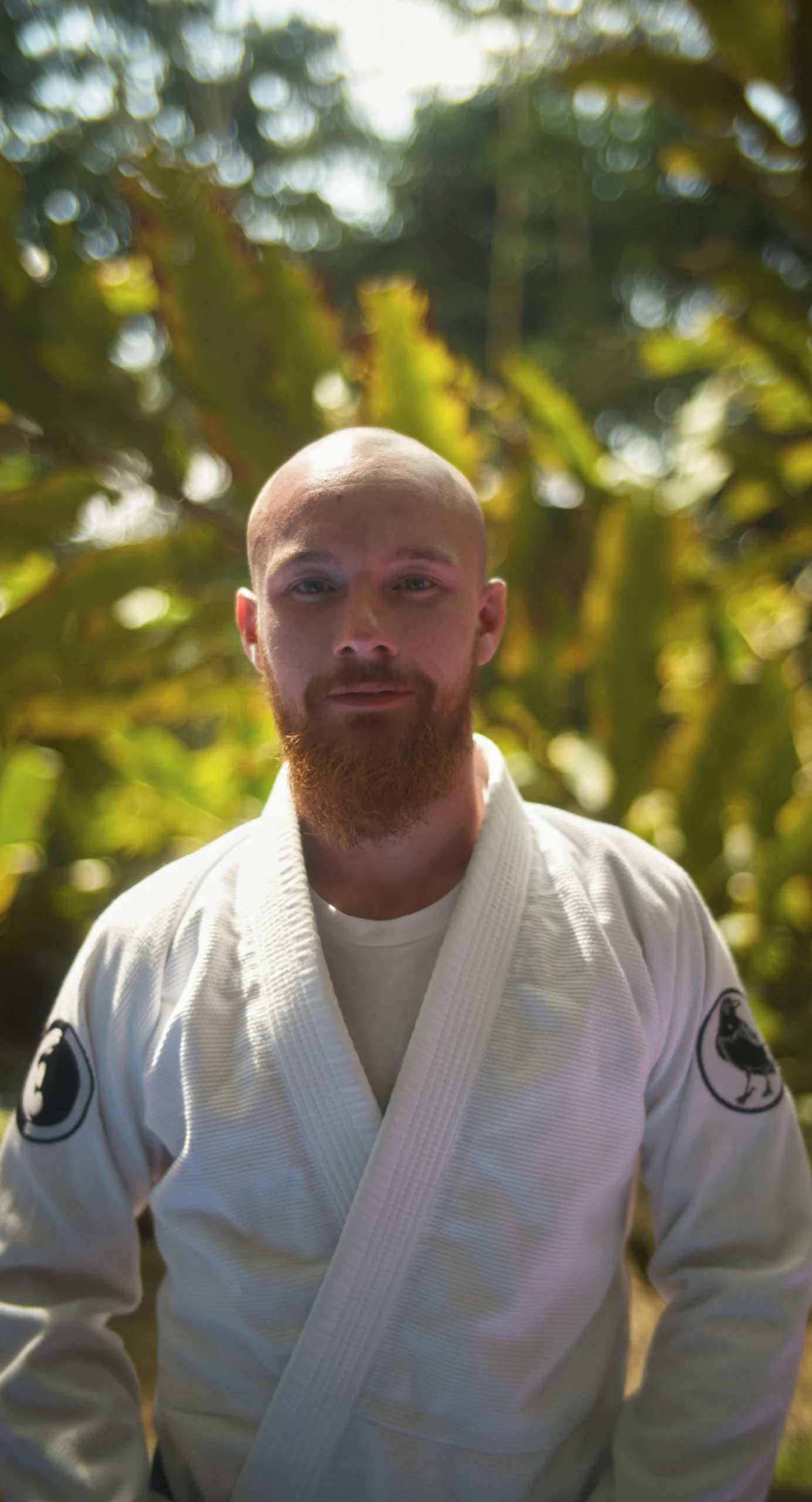 Instructor in a white gi with a beard, smiling and standing in a lush green setting at BJJ Camp XP, showcasing his dedication to teaching Brazilian Jiu-Jitsu.