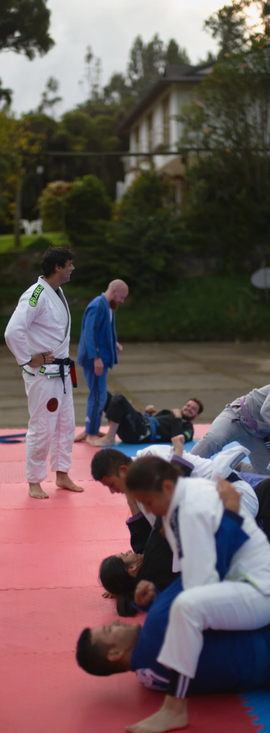 An instructor in a white gi oversees students practicing a Jiu-Jitsu technique at BJJ Camp XP. Participants are performing drills on mats outdoors, with the instructor providing guidance to ensure correct form and technique.