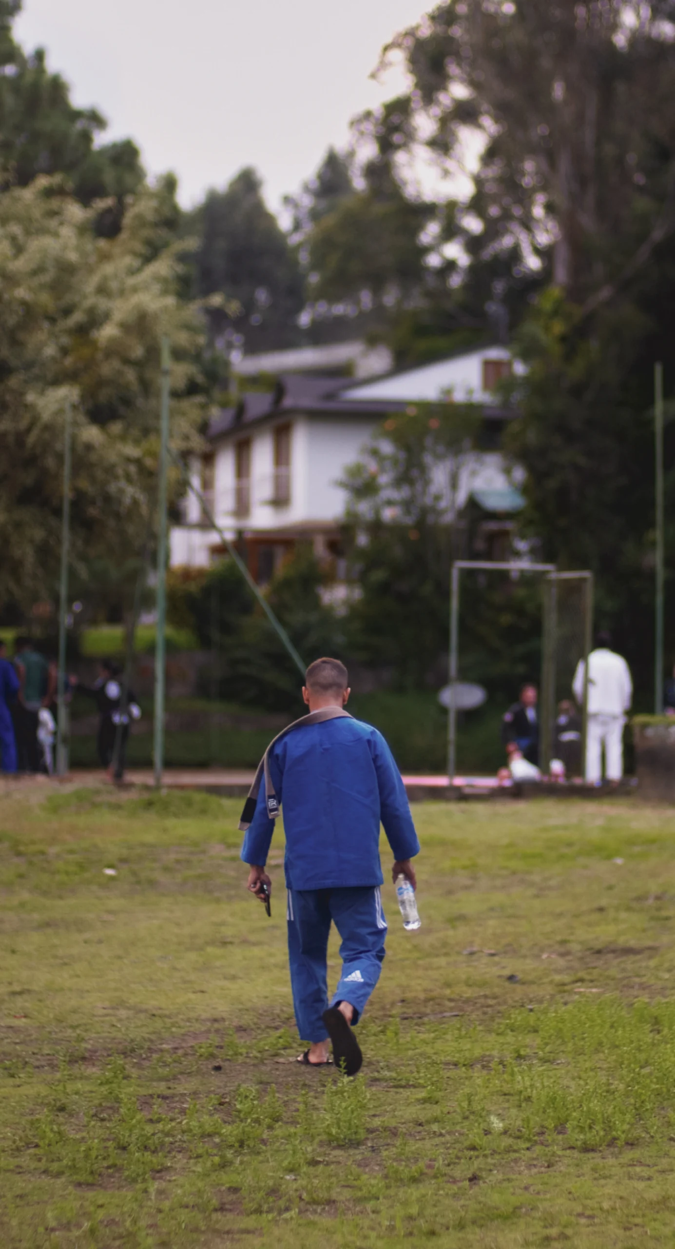 Instructor in a blue gi walking toward the training area at BJJ Camp XP, ready to lead a Jiu-Jitsu session, with a water bottle in hand and a focus on guiding participants in their training amidst a scenic natural backdrop.