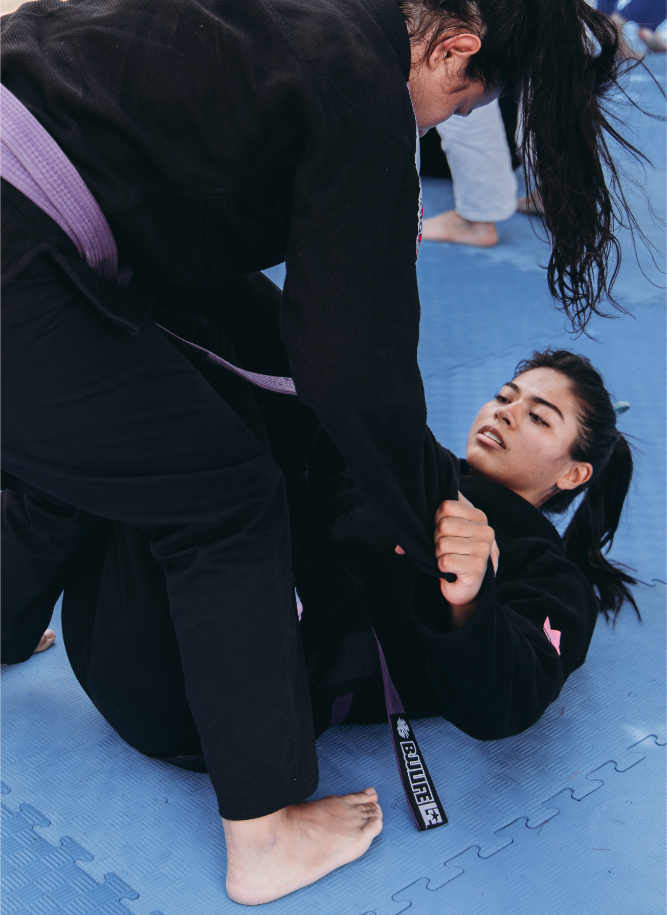 Camila, a member of the XP Athlete Program, practicing Jiu-Jitsu on the mat with a training partner in black gis and purple belts.