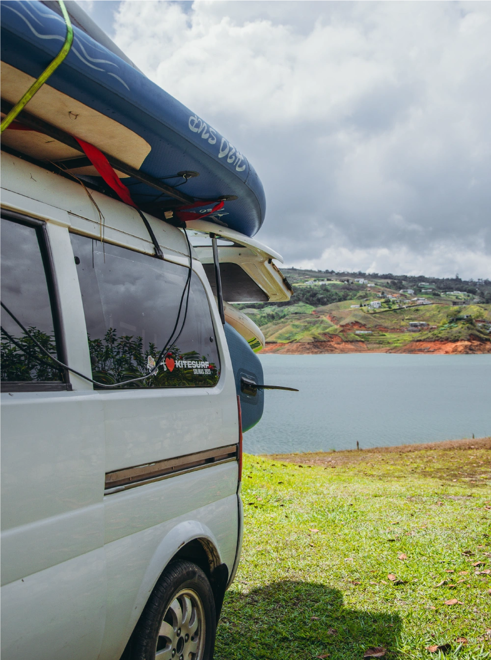 Van packed with paddleboards and kitesurfing gear parked by a scenic lake at a Brazilian Jiu-Jitsu training camp in Colombia.